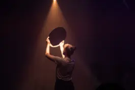 Performer in a theater in a dark space holding a mirror that is relfecting light 