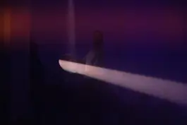 Performer in a theater in a dark space holding a mirror that is relfecting light 